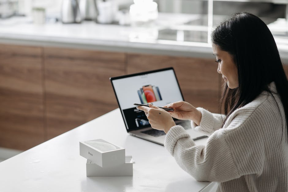 Mujer asiática joven configura un nuevo teléfono inteligente mientras trabaja con la computadora en un interior moderno.