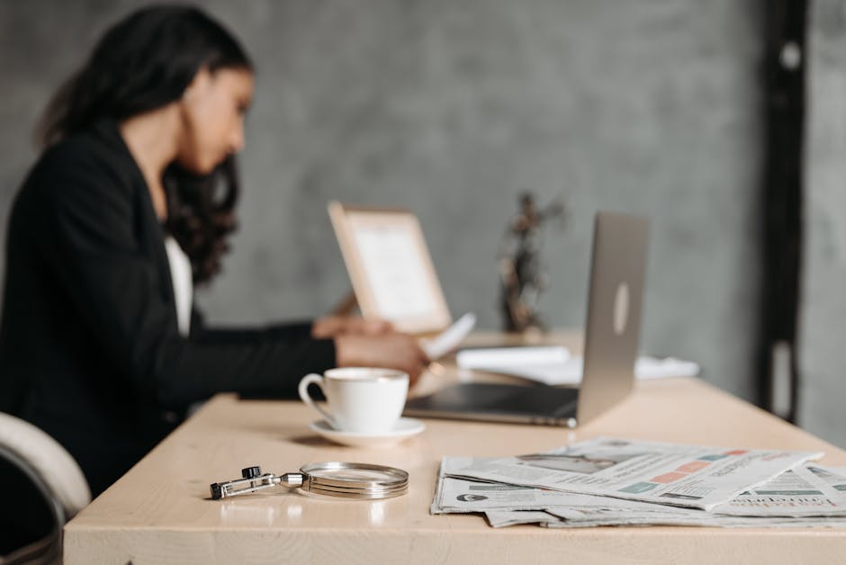 A woman in a professional suit working at a desk with a laptop and documents in an office.