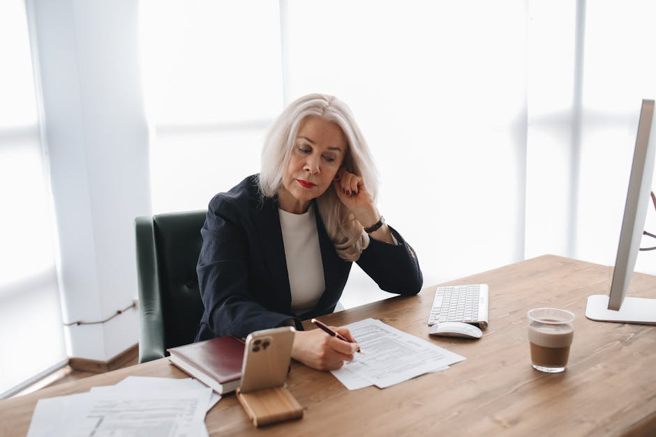 Femme examinant des documents sur un bureau avec un smartphone et un ordinateur dans un bureau moderne.
