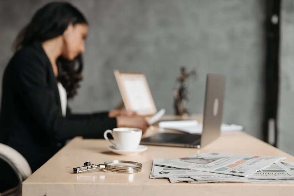 A woman in a professional suit working at a desk with a laptop and documents in an office.