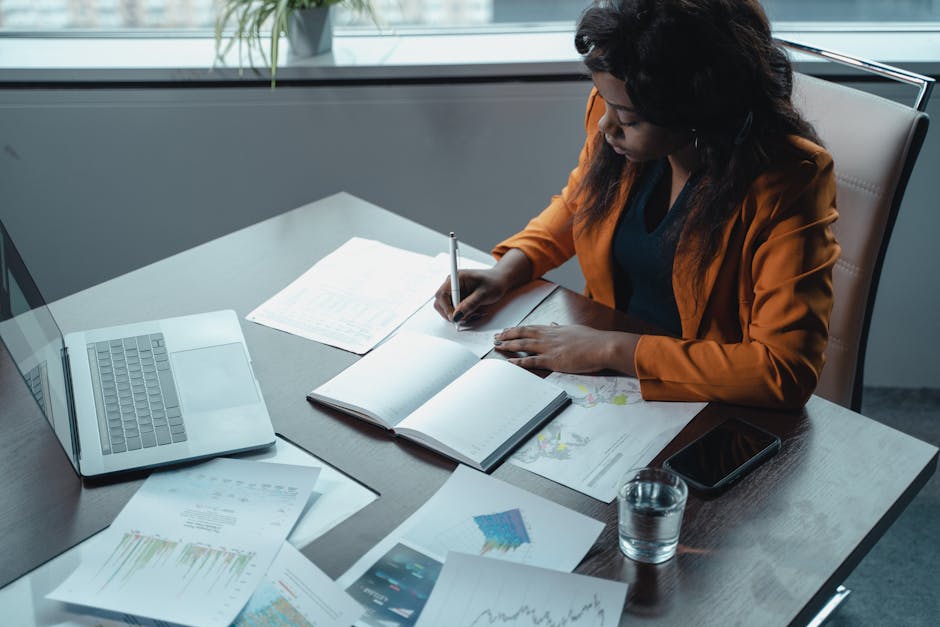 Businesswoman in office writing notes with documents and laptop, focused on analysis and productivity.