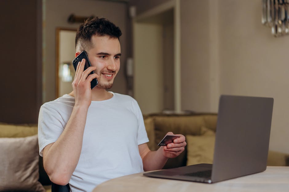 Un jeune homme sourit en faisant du shopping en ligne avec son téléphone et un ordinateur portable, une carte bancaire à la main.