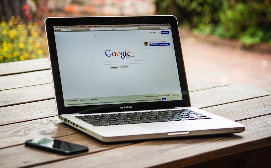 Un MacBook Pro affichant une recherche Google sur une table en bois à l'extérieur, à côté d'un smartphone.